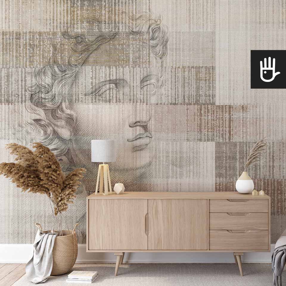 Ścienna tapeta z rysunkiem rzeźby Apolla na jasnym, beżowo-brązowym tle w pionowe prążki, która stanowi tło dla drewnianej komody i dekoracji w stylu boho.