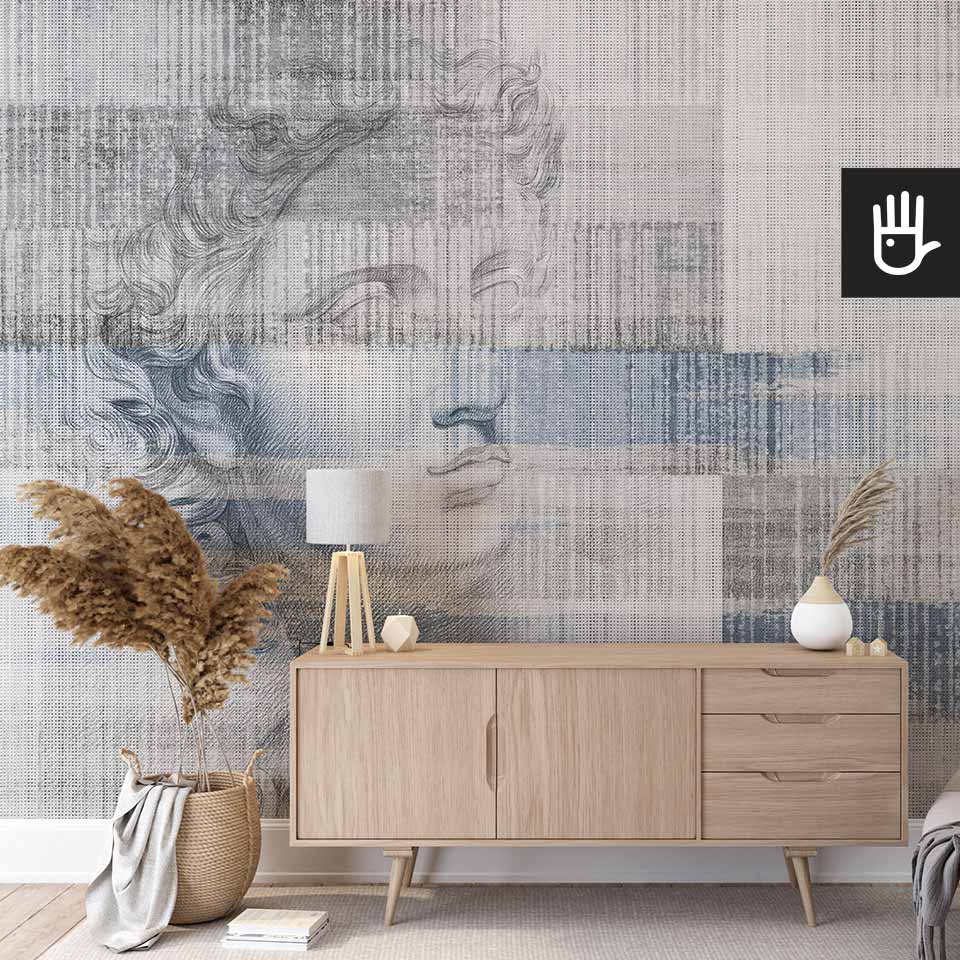 Ścienna tapeta z rysunkiem rzeźby Apolla na jasnym, szaro-niebieskim tle w pionowe prążki, która stanowi tło dla drewnianej komody i dekoracji w stylu boho.